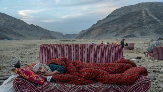 An old woman lies on a sofa in a stony landscape with mountains in the background.