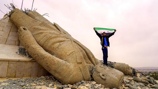 رجل يرفع علم سوريا الحرة واقفًا على رأس تمثال أسد مقلوب.