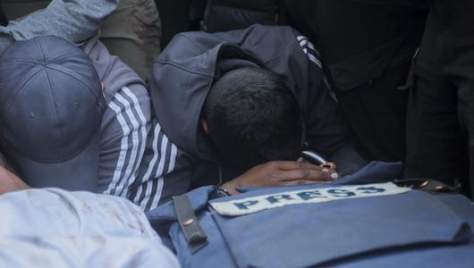 Two men kneel and bow their heads over a blue press vest.