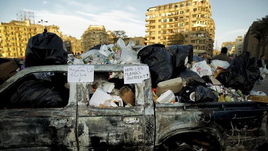 A burnt out truck full of trash bags. On the window frame a sign reads "Here lies Mubark's government!"