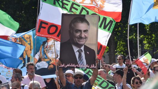 A man in a crowd holds up an image of Crown Prince Reza Pahlavi. There are many flags in the background, including the flag of Iran.