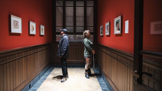 18th Istanbul Biennial (Photo: IKSV | Mete Kaan Özdilek) Two people in a corridor, facing away from eachother and looking at paintings hanging on the walls.