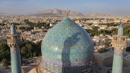 منظر لمسجد الشيخ لطفي الله في أصفهان، إيران. (Photo: Picture Alliance / Amazing Aerial Agency | Airpano) قبة مسجد مزينة بفسيفساء خضراء أمام بانوراما مدينة، وخلفها جبال.