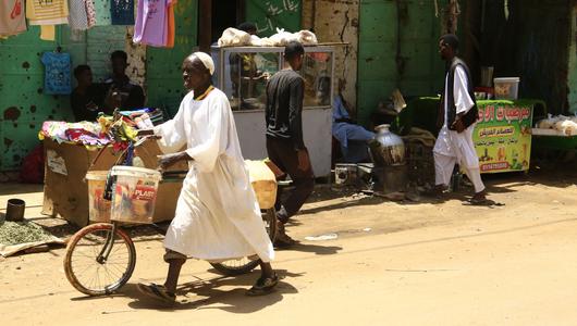 A man pushes his bicycle along a sandy path. In the background a green wall, several people walking and clothes being hung up to dry.