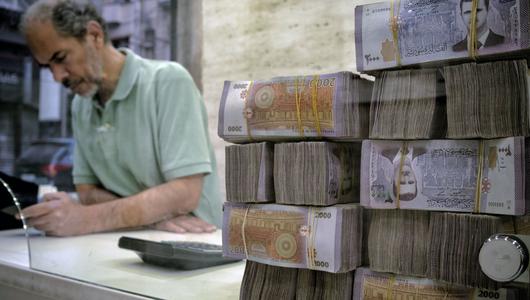 A man stands at a desk looking at his phone. In the foreground, a stack of 2000 SYP notes bearing the face of Bashar al-Assad.