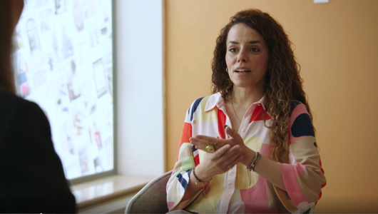 Eine Frau in bunter Bluse sitzt vor einem Fenster und spricht mit ihrem Gegenüber.