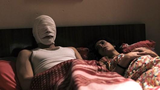 Ein Paar liegt im Bett, der Mann trägt eine Bandage um den Kopf und das Gesicht.