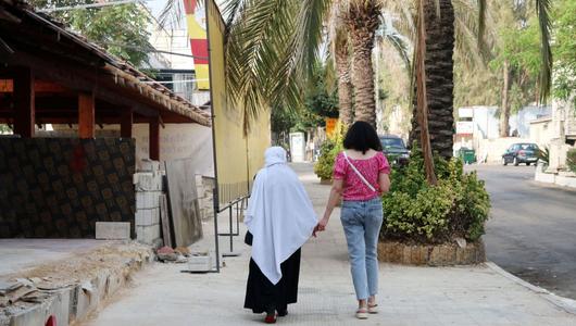 Two women walk down a wide pavement holding hands. One is wearing a headscarf, the other without.