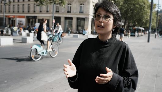 Shahrbanoo Sadat gestikuliert. Im Hintergrund eine Straßenszene, links im BIld eine Fahradfahrerin. 