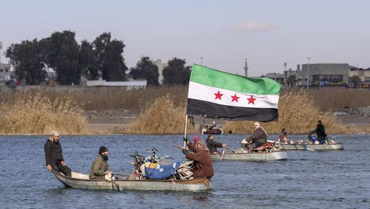 Ein Boot fährt über einen Fluss. Eine Passagierin hält die syrische Nationalflagge in den Händen. 