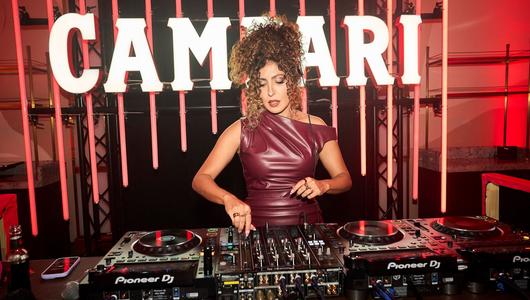 Eine Frau in dunkelrotem Kleid und hochgesteckten Haaren steht am DJ Pult. Hinter ihr Leuchtreklame.