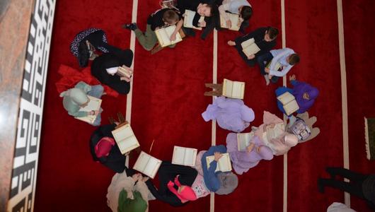 A group of children sit in a circle on a red carpet reading from large books.