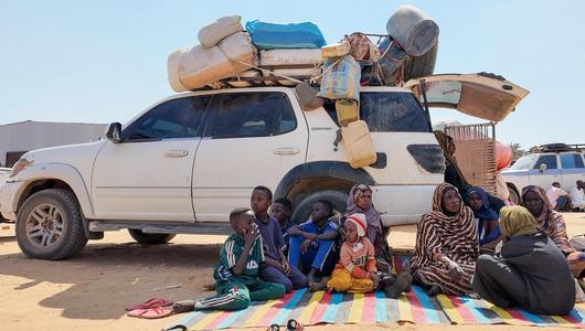 A family sit on a blanket on the ground in front of a truck loaded with bags and suitcases.