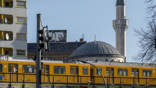 Stadtbild, im Vordergrund fährt eine gelbe Bahn, im Hintergrund ein Wohngebäude und eine Moschee.