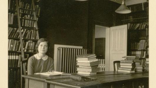 A black and white image of a young woman sitting in a room full of books.