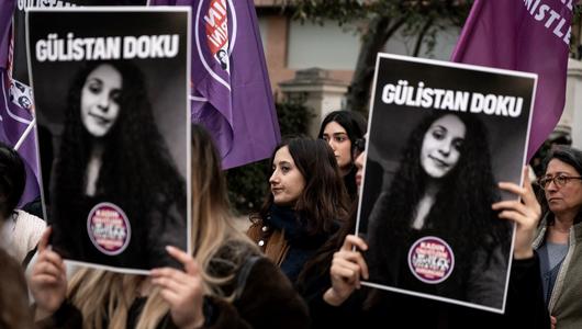 Women are holding up a black-and-white photograph of a young woman, with purple flags in the background.