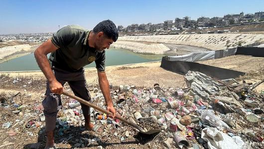 Ein Mann mit kurzem Bart hält eine Schaufel in der Hand; im Blickfeld sind ein Abwasserbasin und einige Zelte zu sehen