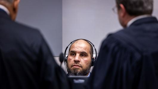 A man is sitting with headphones on; in front of him stand two men in lawyers’ robes, with their backs to the camera.