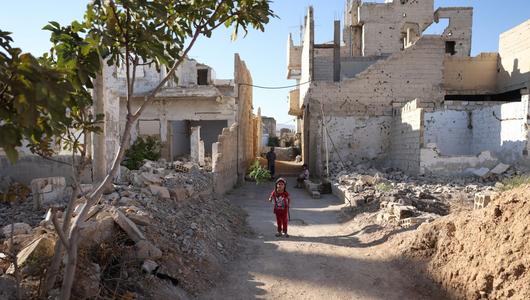 A girl is standing on a path between dilapidated buildings and rubble. In the background are another girl and an old man.