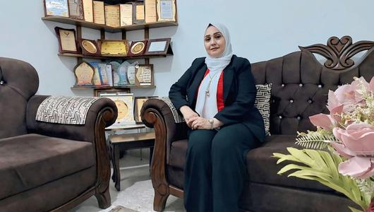 سيدة ترتدي الحجاب وتجلس على أريكة وخلفها رف من الدروع.
