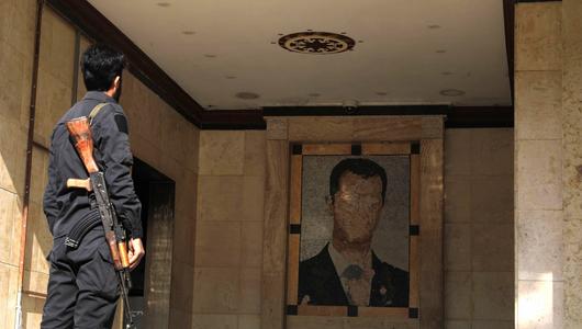 A man in uniform, holding a rifle, seen from behind. He is looking at a damaged mural of Bashar al-Assad.