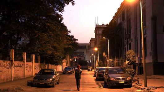 امرأة تسير في طريق خالٍ في جنح الظلام، تضيئه كشافات الإنارة، فيما تصطف السيارات على جانبيه.