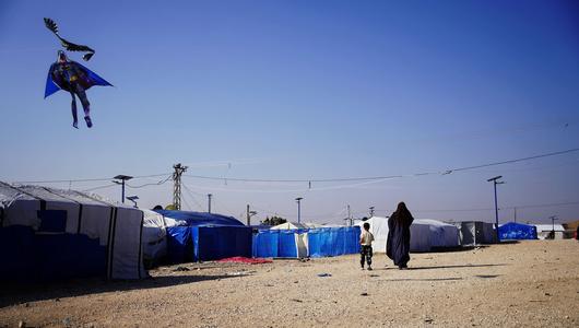 A woman and a child walk towards a cluster of tents. A Batman shaped kite is visible in the sky.